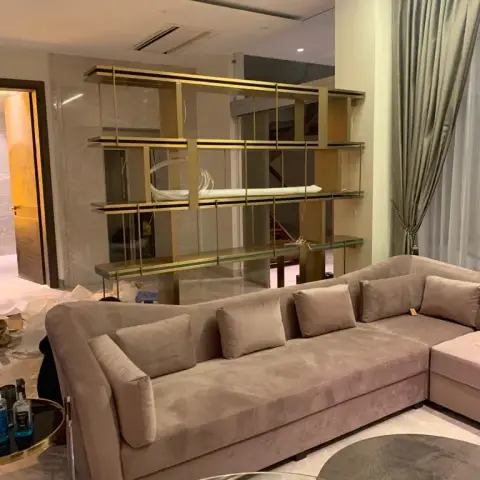 Living room with a brass-framed glass display shelf room divider, a taupe velvet L-shaped sofa, and floor-length sage curtains
