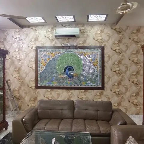 Living room with a brown leather sofa set and a glass-top coffee table beneath a framed peacock mosaic wall art on patterned gold wallpaper.