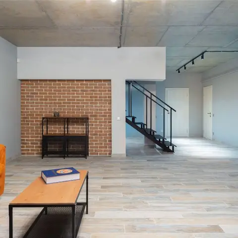 Living room with a burnt-orange sofa against an exposed brick accent wall, raw concrete ceiling, and an open metal staircase behind.