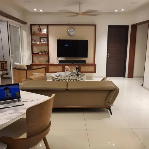 Living room with a cane-fronted TV console and matching cane armchair, a tan leather sofa on tapered wood legs, and a patterned round pouf as coffee table.
