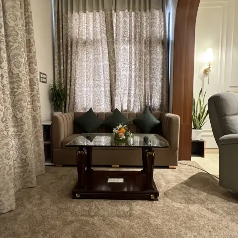 Living room with a carved dark wood and glass coffee table, deep green cushions on a beige sofa, and floor-to-ceiling damask curtains.