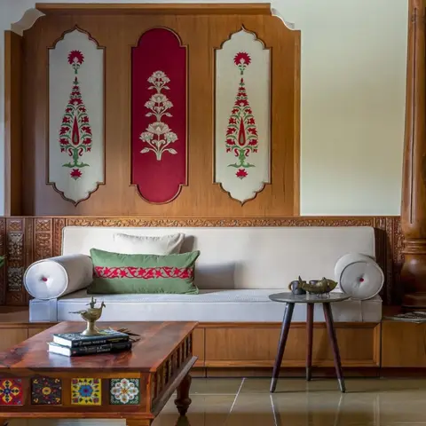 Living room with a carved teak accent wall featuring inset floral tapestry panels, a cream bolster-cushioned daybed sofa, and a hand-painted tile coffee table.