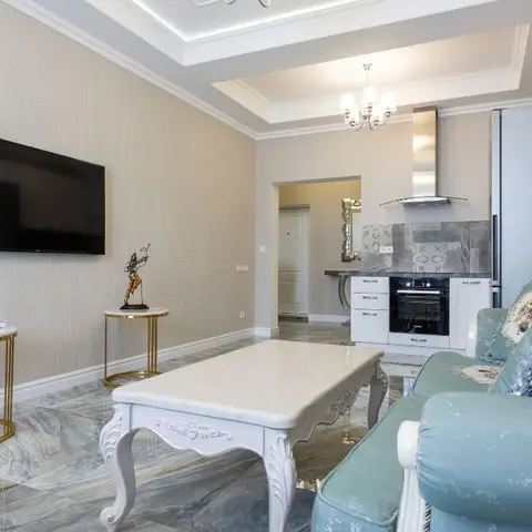 Living room with a carved white coffee table and teal brocade sofa on marble flooring, opening into the kitchen beyond under a cove-lit ceiling.