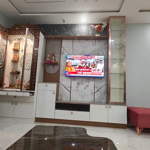 Living room with a carved wood pooja unit with fretwork jaali doors beside a TV console featuring a marble-pattern backsplash and rose-gold trim