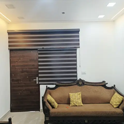Living room with a carved wood-frame sofa upholstered in brown with yellow patterned cushions, zebra blinds on the window, and cove ceiling lighting.