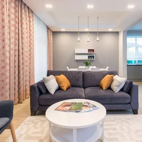 Living room with a charcoal-blue sofa and mustard accent cushions on a patterned area rug, floral-patterned curtains, and a round white coffee table