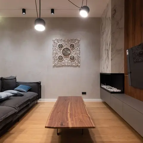 Living room with a charcoal sofa facing a live-edge walnut coffee table, a carved wood-and-metal wall clock on a plaster wall