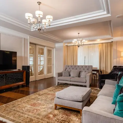 Living room with a coffered crown-moulded ceiling, twin crystal chandeliers, a tufted grey loveseat and sofa with teal accent cushions on a Persian-style rug