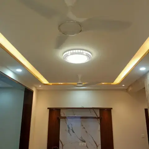 Living room with a cove-lit false ceiling holding two fans and a round chandelier-style light above a marble-backed TV unit wall flanked by dark wood panels.