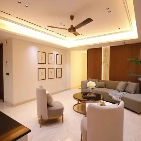 Living room with a cove-lit ceiling fan, cream armchairs with gold legs and a grey sectional sofa around round nesting coffee tables, a six-frame gallery wall