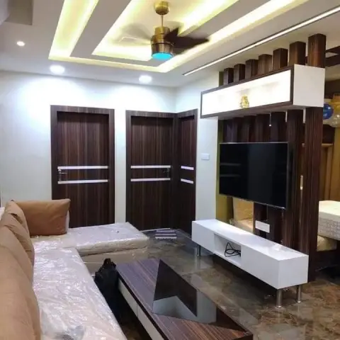 Living room with a cove-lit stepped false ceiling, a veneer TV unit beside a fluted wood partition, and three matching wooden internal doors
