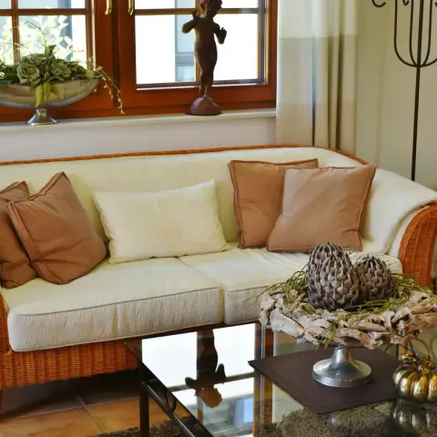 Living room with a cream wicker sofa set with tan cushions, a glass-top coffee table, and terracotta floor tiles by a wood-framed window.