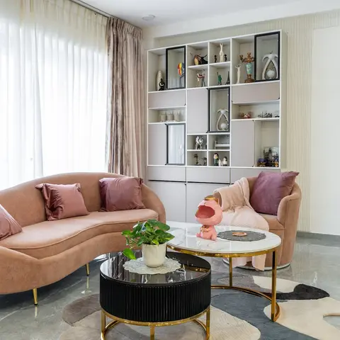 Living room with a curved blush-pink velvet sofa, a mauve armchair, twin gold-framed coffee tables, and a backlit display unit styled with curios.