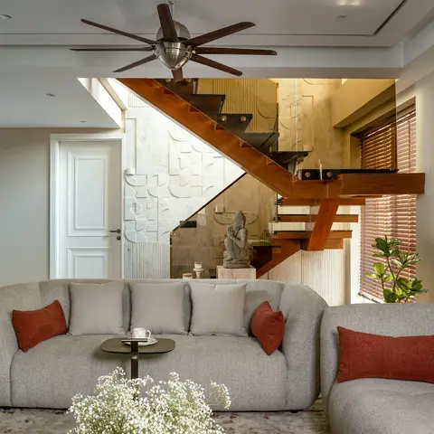 Living room with a curved boucle sofa in grey and rust cushions, a wood-and-glass staircase behind, and a large 3D-textured stone accent wall.