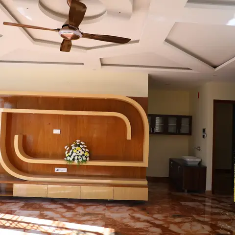 Living room with a curved two-tone wood TV display unit, a geometric cove-lit ceiling fan, and a carved wooden main door dressed with marigold garlands.