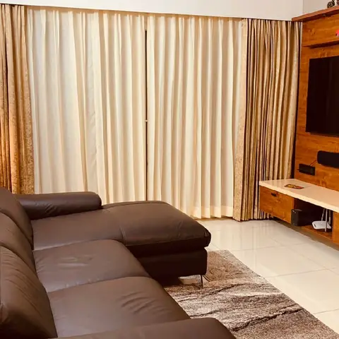 Living room with a dark brown leather L-shaped sofa facing a wood-panelled TV wall with a floating console, floor-length curtains and a patterned area rug.