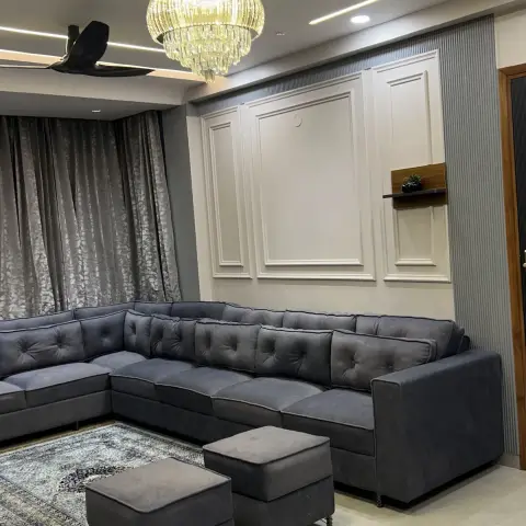 Living room with a dark grey tufted L-shaped sectional sofa and matching ottomans, under a tiered crystal chandelier and a raised-panel moulded wall.