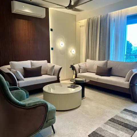 Living room with a dark wood-panelled accent wall, sculptural arched wall sconces, a ribbed round coffee table between beige sofas and a teal velvet tub chair.