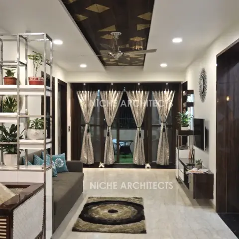 Living room with a dark-wood diamond-inlay false ceiling, grey sofa, metal etagere styled with potted plants, and sheer-curtained glass doors to a balcony.