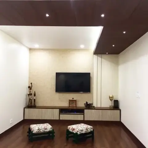 Living room with a dark wood panelled ceiling and cove lighting over a beige textured-wallpaper TV wall, flanked by two floral-pattern wooden stools.