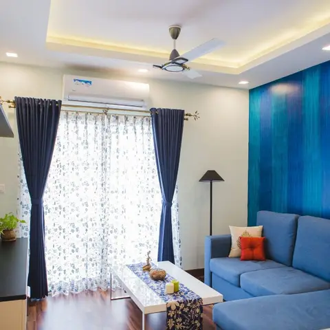 Living room with a deep blue textured accent wall behind a navy sectional sofa, layered navy-and-floral sheer curtains, a cove-lit ceiling with fan