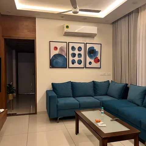 Living room with a deep teal L-shaped sofa, a three-panel abstract art gallery wall, a wood-and-glass coffee table, and a cove-lit ceiling.