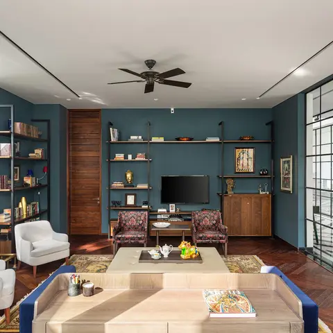 Living room with a deep teal accent wall behind an open metal-and-wood shelving/TV unit, patterned armchairs, a herringbone wood floor, and a marble fireplace.