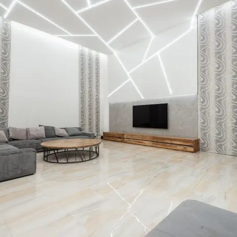 Living room with a dramatic angular LED cove ceiling, a grey sectional sofa, a round wood coffee table and swirl-pattern drapery on marble-look flooring