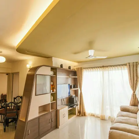 Living room with a dramatic curved cove-lit ceiling bulkhead above a matching curved wood TV and display unit, sheer curtains framing a bright balcony door.