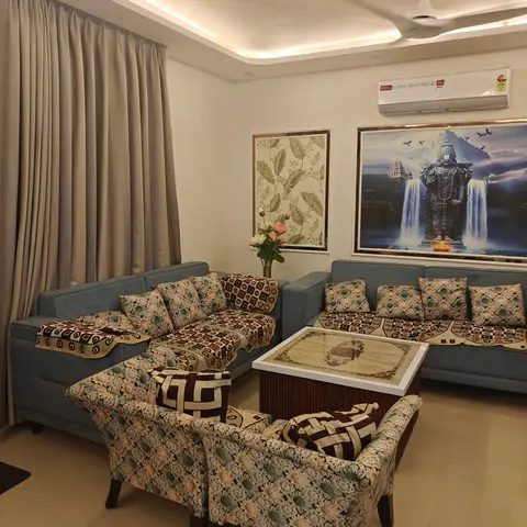 Living room with a dusty-blue L-shaped sofa and matching floral-upholstered armchairs, a religious wall painting, and a cove-lit ceiling border.