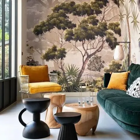Living room with a floor-to-ceiling tree mural wallpaper, an emerald velvet sofa, a mustard velvet armchair, and sculptural black and wood side tables.