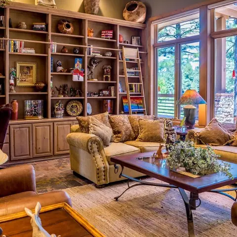 Living room with a full-wall built-in bookshelf styled with southwestern curios, a leather-and-fabric sectional, and floor-to-ceiling windows to a wooded view.