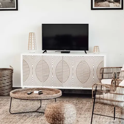 Living room with a geometric carved-pattern white and wood TV console, rattan armchairs, a round rattan-top coffee table, and a jute rug