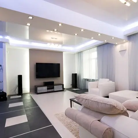Living room with a glossy cove-lit false ceiling, a wall-panelled TV console flanked by floor speakers, checkerboard floor tiles