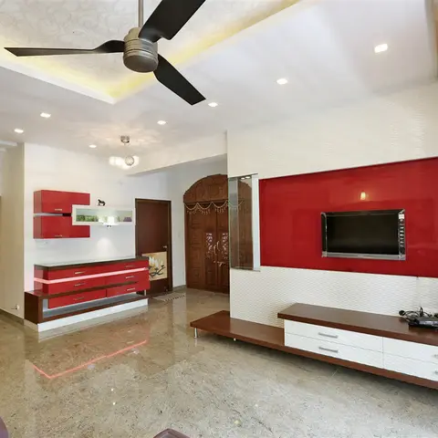 Living room with a glossy red TV feature wall and matching red overhead display unit, marble flooring, and a carved wooden pooja door visible in the background.