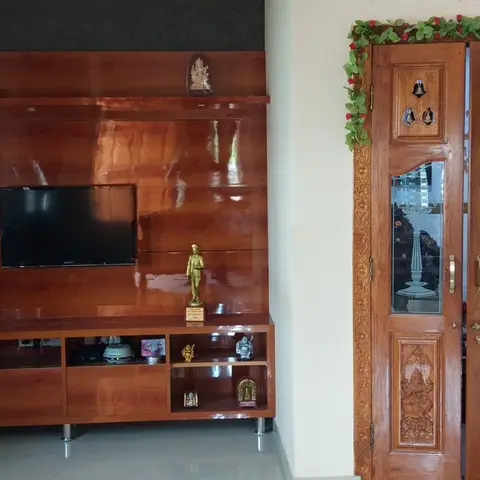 Living room with a glossy wood-veneer TV unit displaying brass idols, beside an ornately carved wooden pooja door with hanging bells and a floral toran.