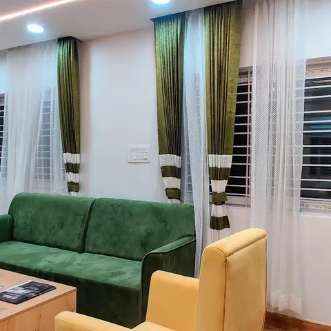 Living room with a green velvet sofa and mustard-yellow armchairs around a wooden coffee table, framed by olive-green striped curtains and a cove-lit ceiling.