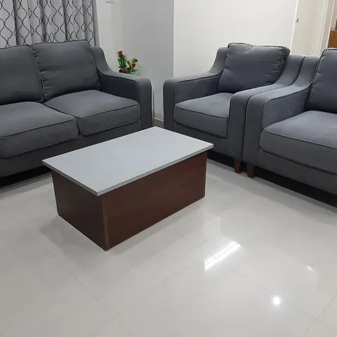 Living room with a grey fabric three-piece sofa set arranged around a wood-and-white coffee table, patterned sheer curtains, and plain white walls and flooring.