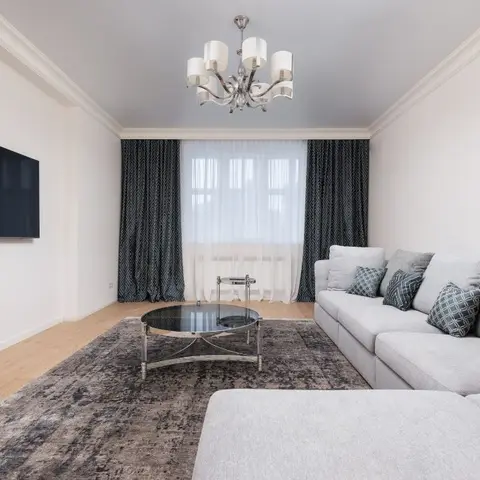 Living room with a grey L-shaped sectional sofa, round glass coffee table, and layered sheer-and-patterned curtains under a crystal chandelier.