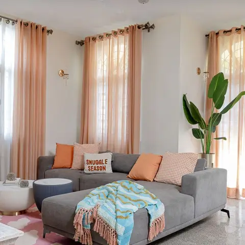 Living room with a grey L-shaped sofa styled with peach and gingham cushions and a blue tasseled throw, peach sheer curtains