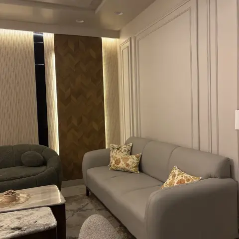 Living room with a grey sofa and green boucle armchair around nested marble coffee tables, a backlit chevron wood accent panel and moulded wall panelling.