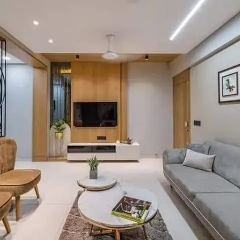 Living room with a grey three-seater facing a TV on an oak-panelled wall, two tan lounge chairs on a dark rug with round marble side tables