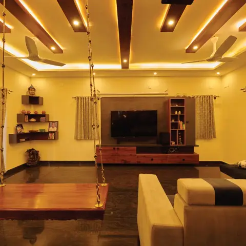 Living room with a hanging wooden swing, exposed wood-beam cove-lit ceiling, and a wood-veneer TV unit against a mustard wall.
