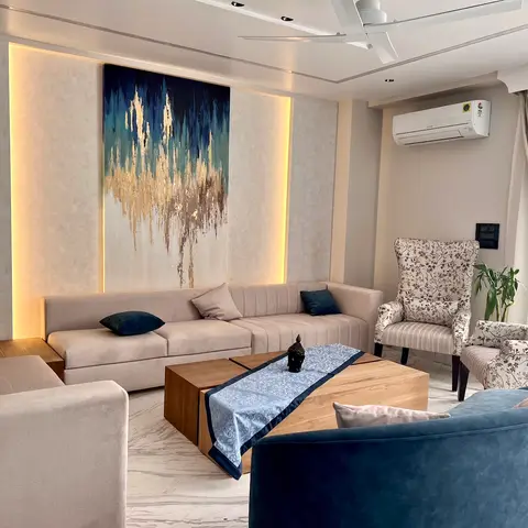 Living room with a large backlit blue and gold abstract canvas above a beige sofa, floral wingback chairs and a blue velvet sofa around a wood coffee table