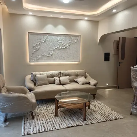 Living room with a large backlit textured art panel above a beige tufted leather sofa and wingback armchair (still in protective wrap) around a round
