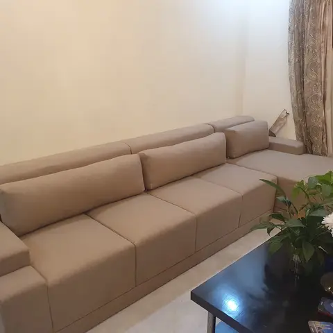 Living room with a large beige L-shaped sectional sofa, a black coffee table styled with fresh flowers, and patterned curtains by the balcony door.