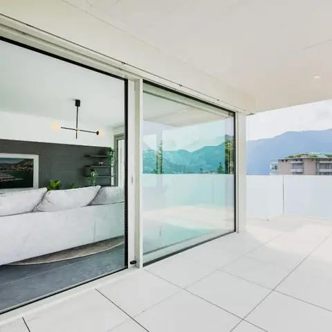 Living room with a light grey sofa against a dark wood-panelled TV wall, opening via full-height sliding glass doors onto a large tiled terrace with mountain