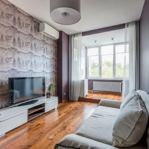 Living room with a lilac forest-print wallpaper feature wall behind a wall-mounted TV on a white and walnut console, sheer curtains framing a window seat