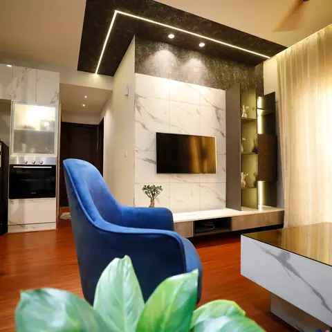 Living room with a marble-clad, cove-lit TV feature wall, a blue velvet armchair, a marble-top coffee table, and sheer gold curtains