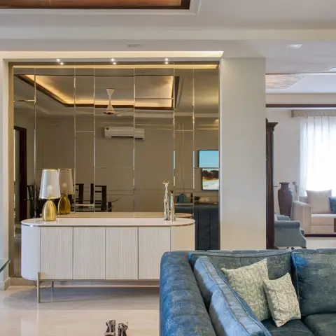 Living room with a mirror-clad wall reflecting the dining area beyond, an oval fluted console with brass lamps, and a deep teal velvet sofa in the foreground.